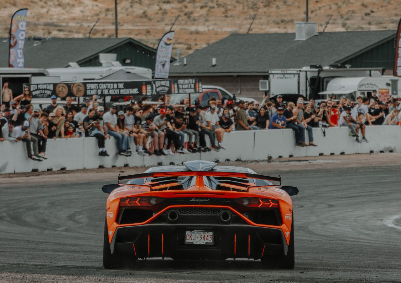 Services-01 A stunning orange Lamborghini Aventador speeds at a race track with spectators in the background.
